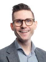 Photo of Brennen Schmidt wearing glasses and blue, white, and black pattern dress shirt with grey suit jacket.